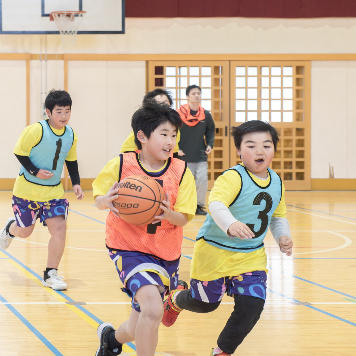 ボールを持って走る子ども3人と、後ろに立つコーチ
